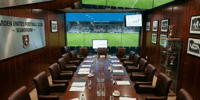 Boardroom meeting of Ardeen United Football Club with executives seated around a long table and stadium visible through large windows.