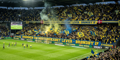 Crowded football stadium with Brøndby IF fans waving yellow and blue flags and smoke flares.